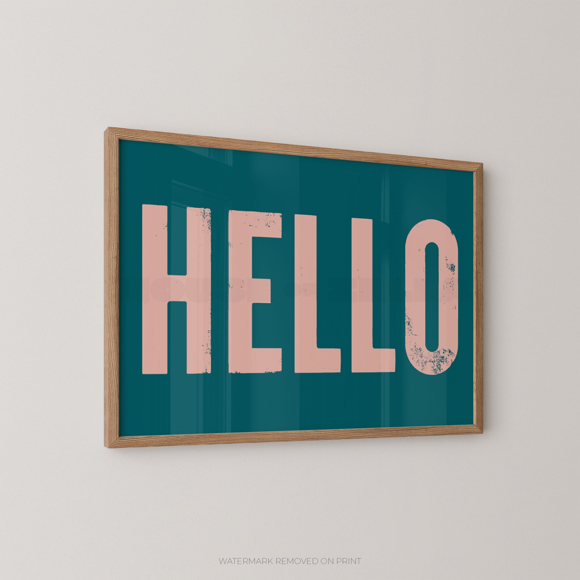 Framed wall art print with the word "HELLO" in large distressed pink letters on a teal background, shown hanging on a light gray wall.