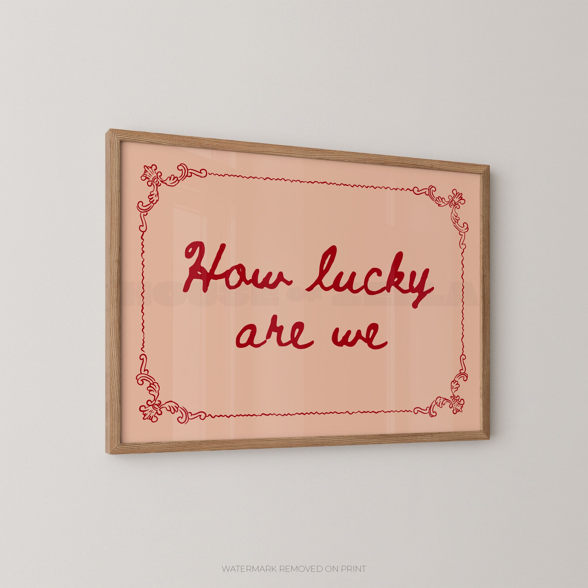 Framed wall art print with the phrase “How lucky are we” in red script on a pale peach background, bordered by a thin red decorative line and a light wood frame, shown against a light gray wall.