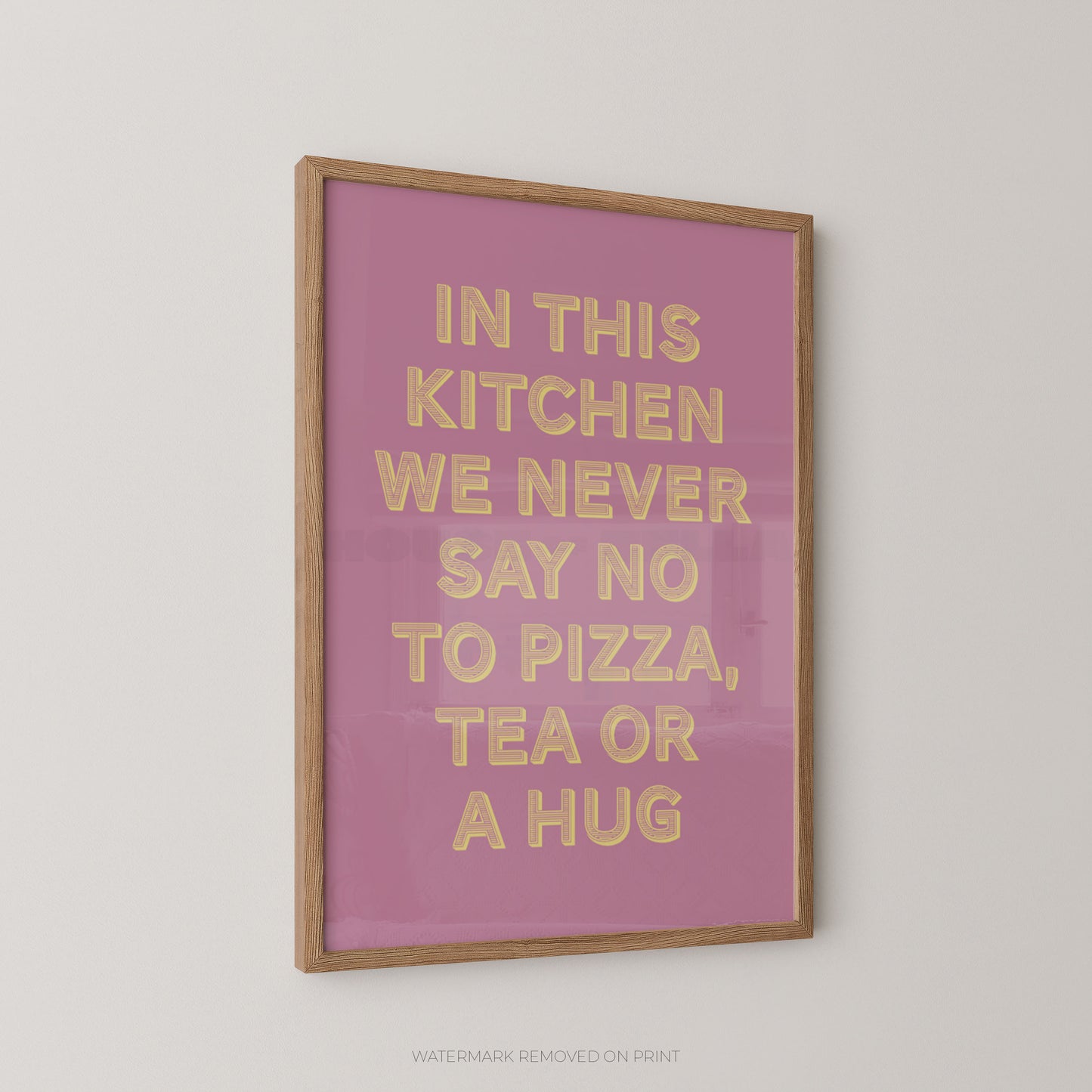 A framed typographic wall art print with a pink background and raised gold lettering that reads, 'IN THIS KITCHEN WE NEVER SAY NO TO PIZZA, TEA OR A HUG,' shown hanging on a light neutral wall.