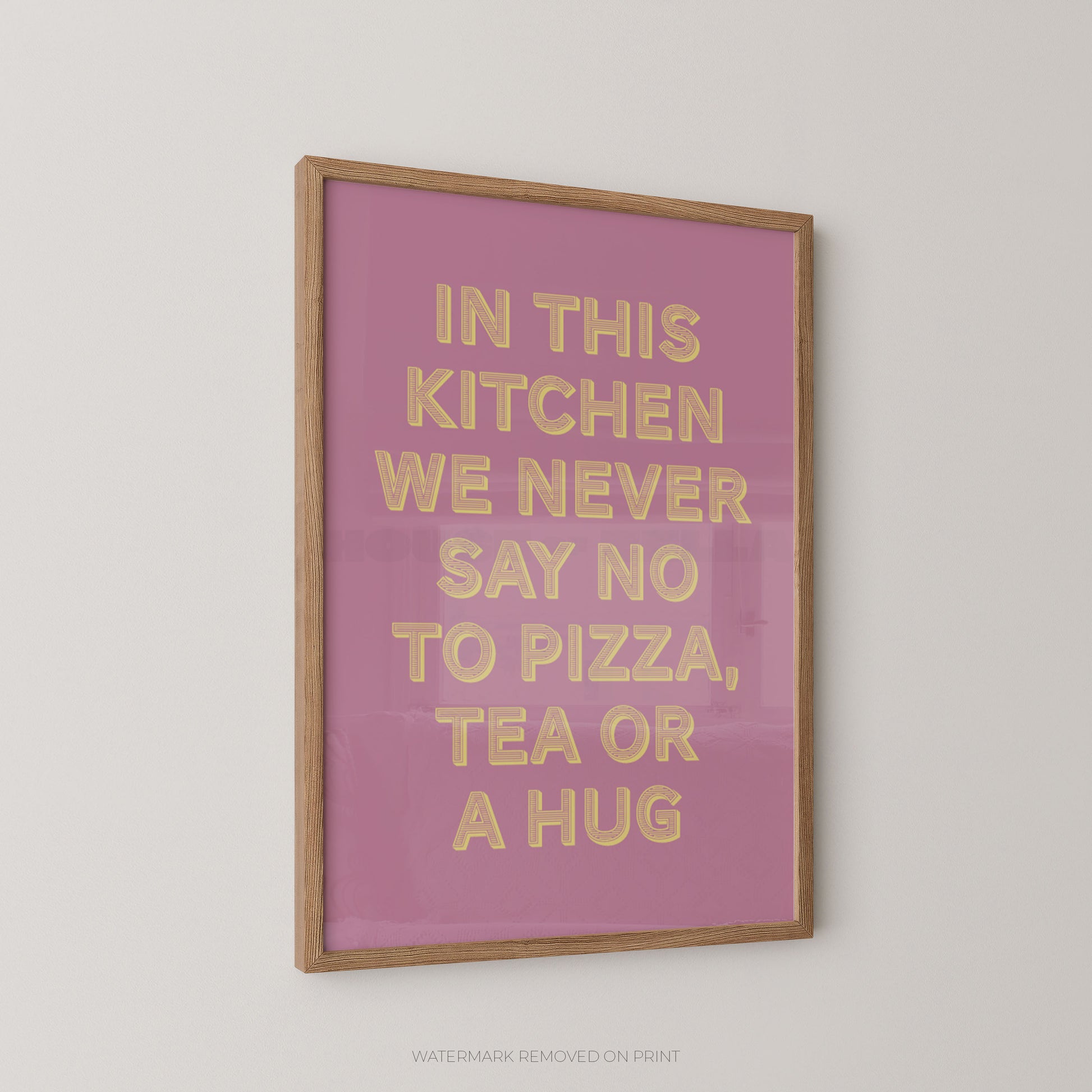 A framed typographic wall art print with a pink background and raised gold lettering that reads, 'IN THIS KITCHEN WE NEVER SAY NO TO PIZZA, TEA OR A HUG,' shown hanging on a light neutral wall.