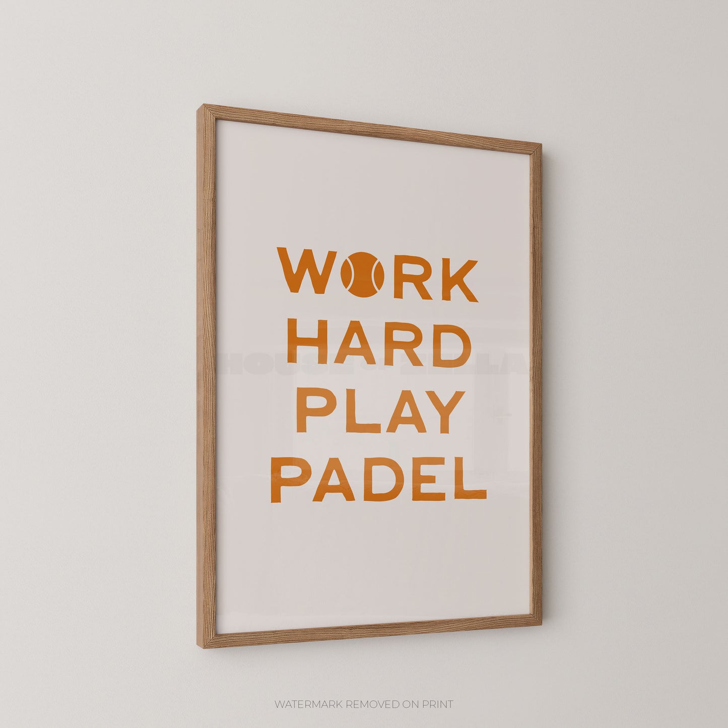 A framed typographic wall print with the phrase “WORK HARD PLAY PADEL” in orange lettering on a white background, shown hanging on a light gray wall. The print has a light wood frame, and a tennis ball icon appears inside the letter O.