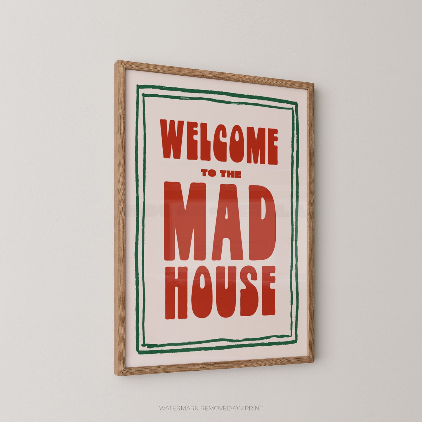 Framed wall art print with the text 'WELCOME TO THE MAD HOUSE' in large red letters, bordered by a green hand-drawn line on an off-white background, shown in a light wood frame against a light gray wall.