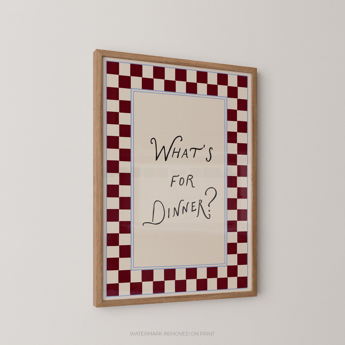 A framed wall art print with the handwritten text 'WHAT'S FOR DINNER?' centered on a cream background, surrounded by a red-and-white checkered border and a light wood frame, shown hanging on a plain light wall.