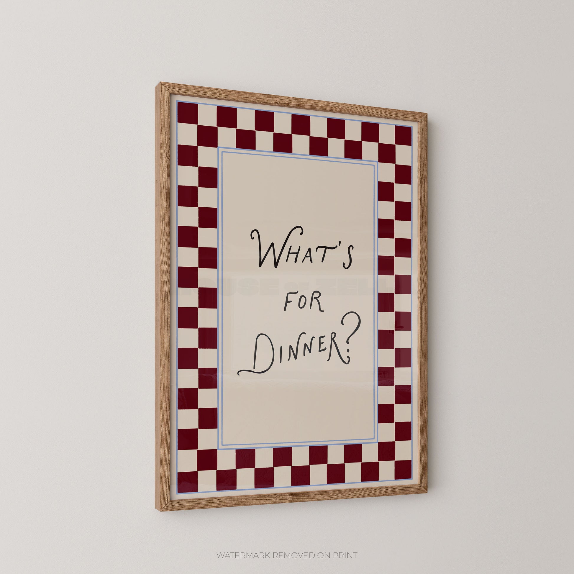 A framed wall art print with the handwritten text 'WHAT'S FOR DINNER?' centered on a cream background, surrounded by a red-and-white checkered border and a light wood frame, shown hanging on a plain light wall.