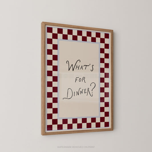 A framed wall art print with the handwritten text 'WHAT'S FOR DINNER?' centered on a cream background, surrounded by a red-and-white checkered border and a light wood frame, shown hanging on a plain light wall.
