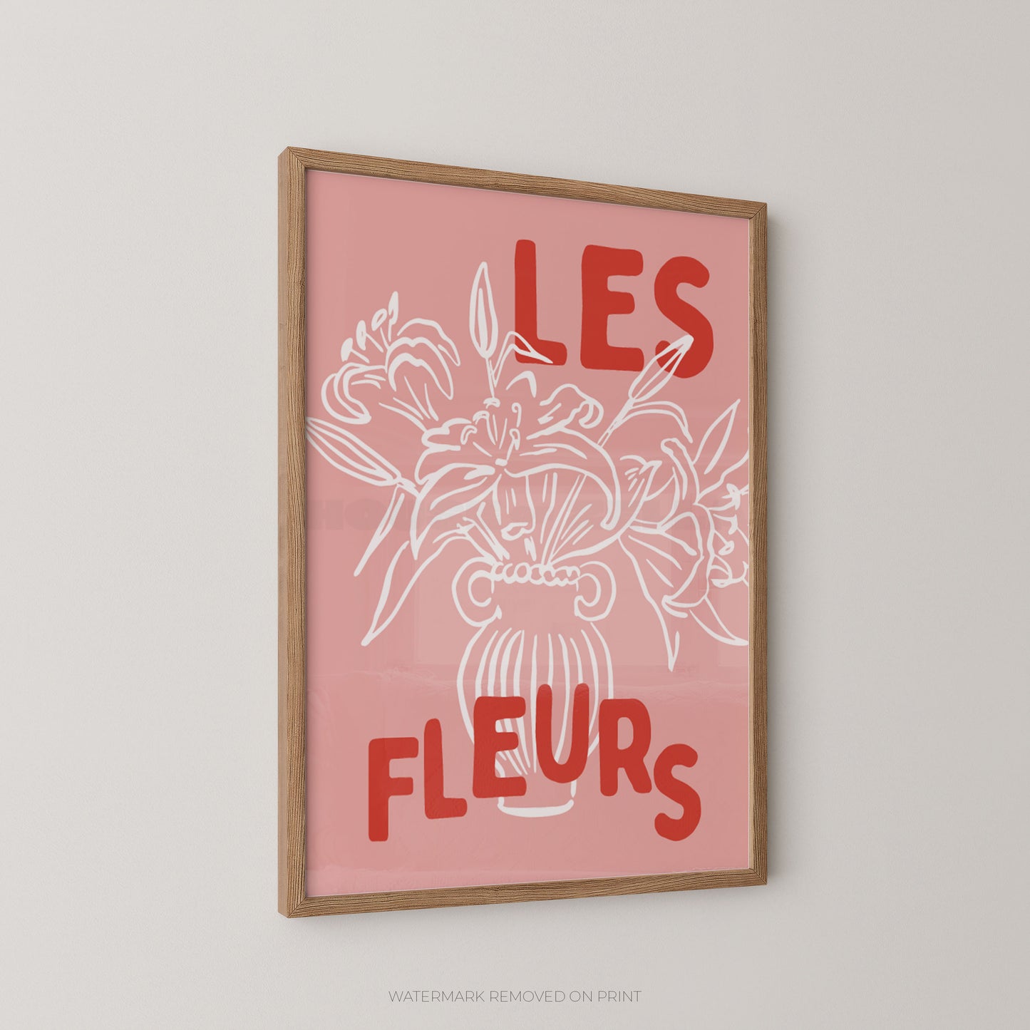 A framed wall art print with a pink background, white line drawing of flowers in a vase, and red text reading “LES FLEURS,” shown hanging on a light gray wall.