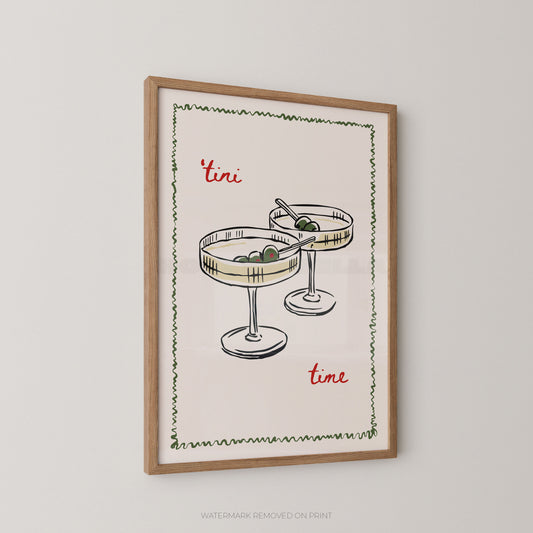 Framed wall art print featuring a hand-drawn illustration of two margarita glasses with olives, bordered by a green zigzag line, with the words "tini time" in red script on a cream background, shown in a light wood frame against a light gray wall.