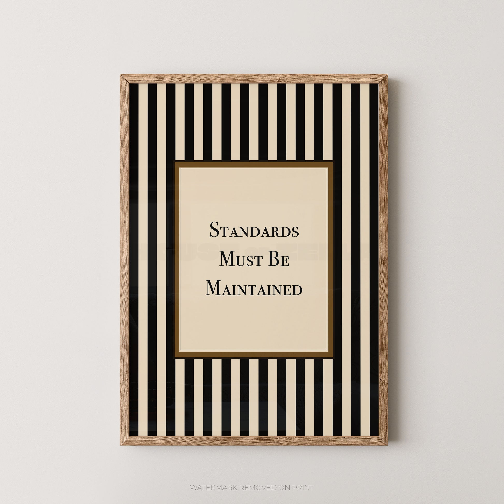 Framed wall art print with vertical black and cream stripes, a central cream panel, and the text 'STANDARDS MUST BE MAINTAINED' in black serif lettering, shown against a light neutral wall.