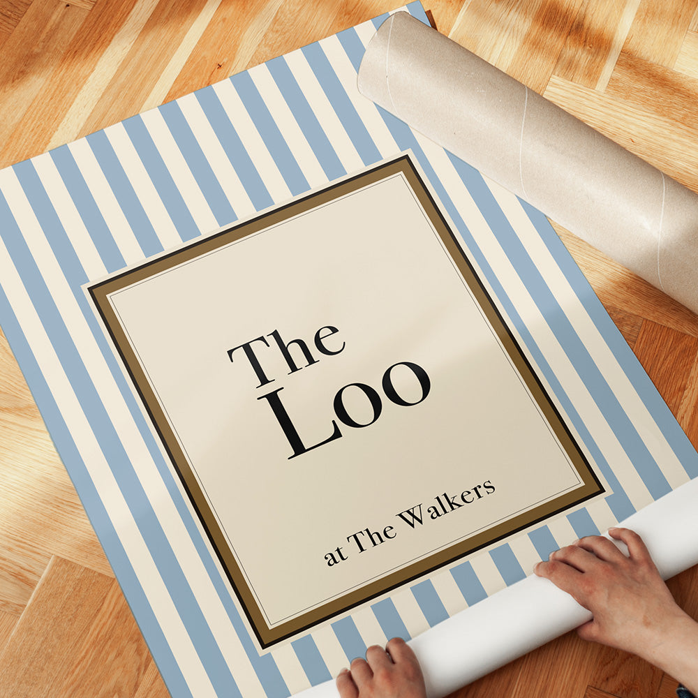 Framed print with 'The Loo at The Walkers' text on a blue and white striped mat, being handled on a wooden floor.