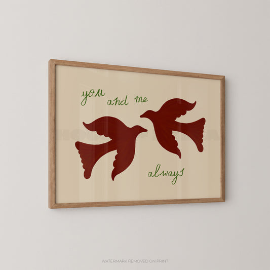 Framed wall art print with two flying birds in dark red on a warm cream background, with the words 'you and me' and 'always' in green script, shown hanging on a light gray wall.