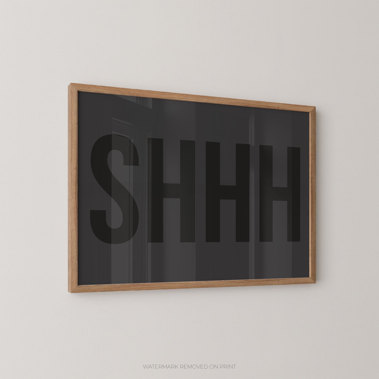Framed wall art featuring the word 'SHHH' in large black letters on a dark charcoal background, enclosed in a light natural wood frame and mounted on a light gray wall.