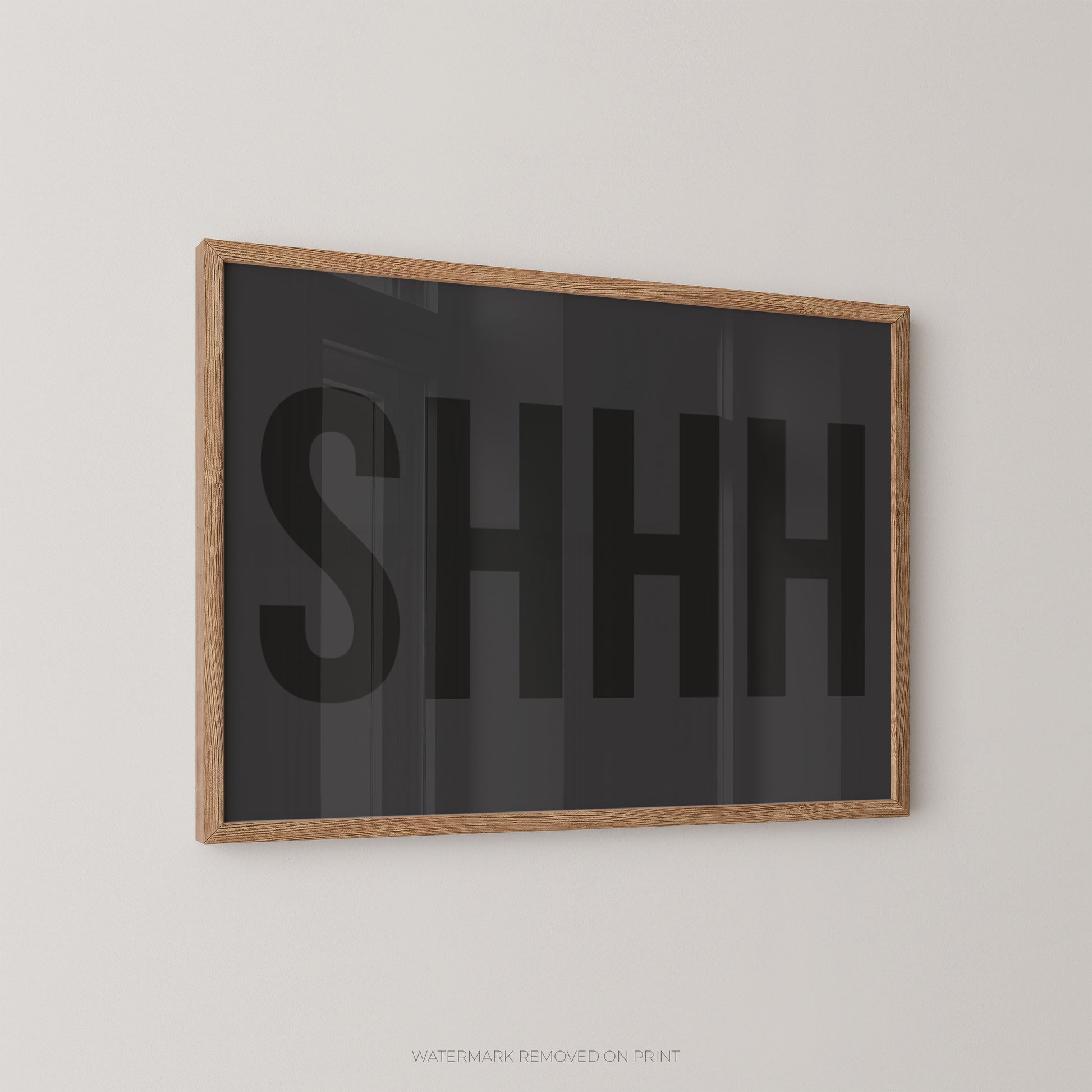 Framed wall art featuring the word 'SHHH' in large black letters on a dark charcoal background, enclosed in a light natural wood frame and mounted on a light gray wall.