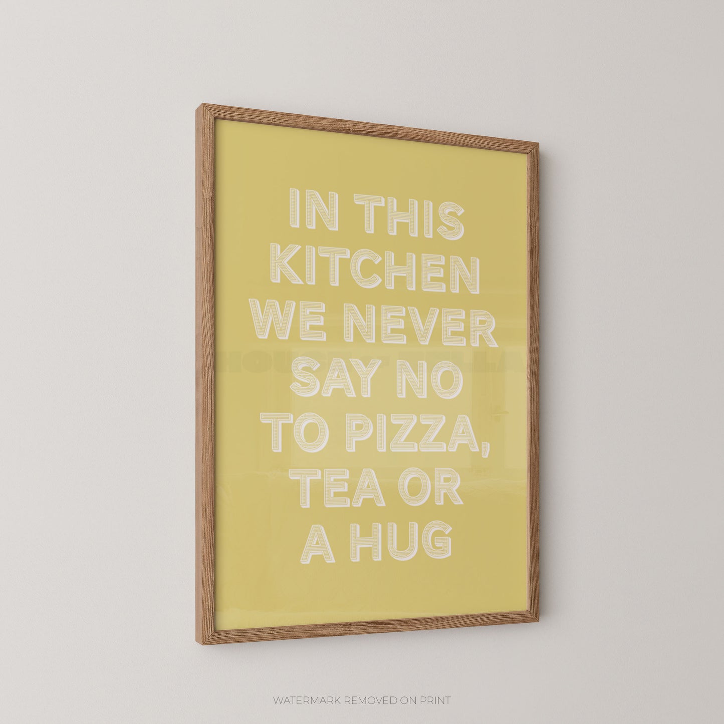 A framed wall art print with a light wood frame and a pale yellow background, featuring raised white text that reads, 'IN THIS KITCHEN WE NEVER SAY NO TO PIZZA, TEA OR A HUG.' The print is shown hanging on a light gray wall.