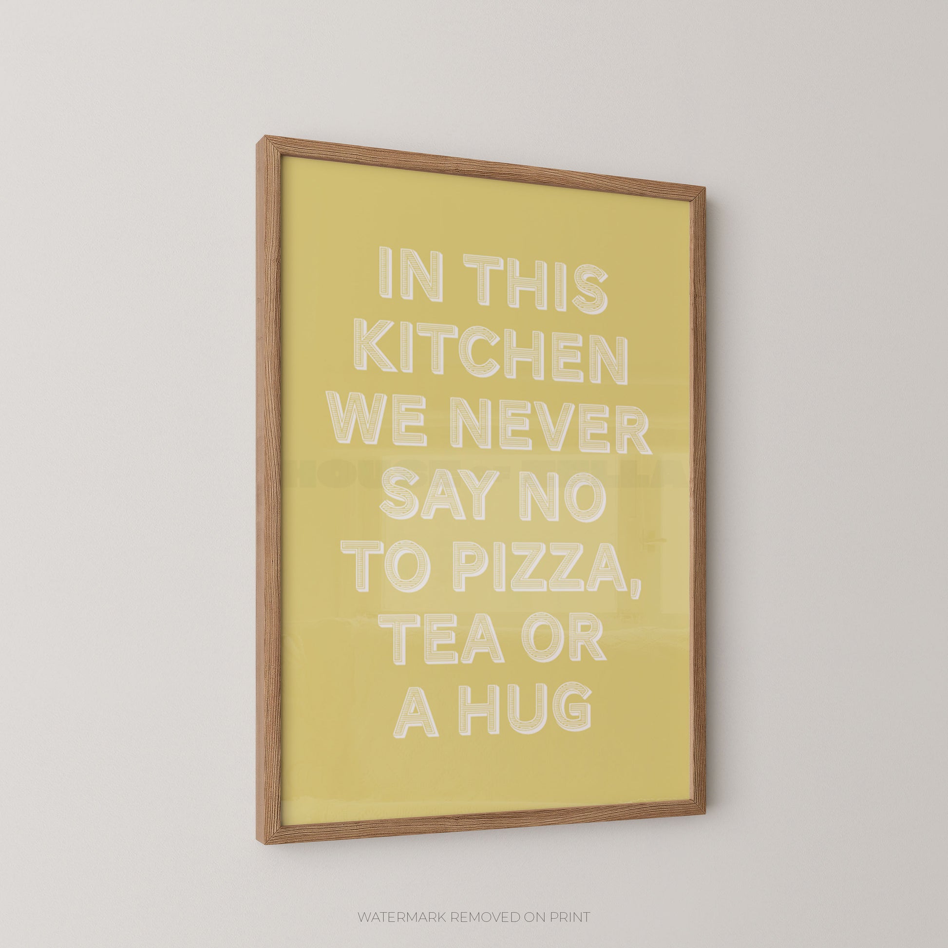 A framed wall art print with a light wood frame and a pale yellow background, featuring raised white text that reads, 'IN THIS KITCHEN WE NEVER SAY NO TO PIZZA, TEA OR A HUG.' The print is shown hanging on a light gray wall.