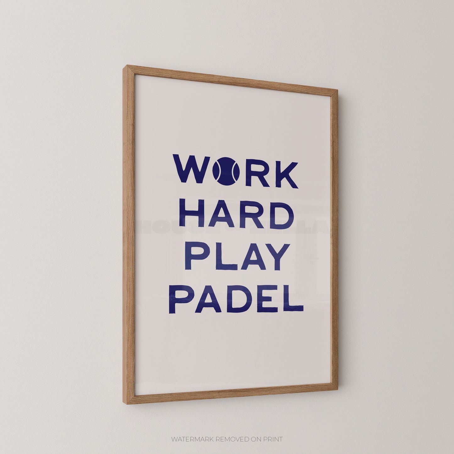 A framed typographic wall print with the phrase “WORK HARD PLAY PADEL” in dark blue text on a white background, shown hanging on a light neutral wall. The print has a light natural wood frame and a small tennis ball icon replacing the letter O in “WORK.