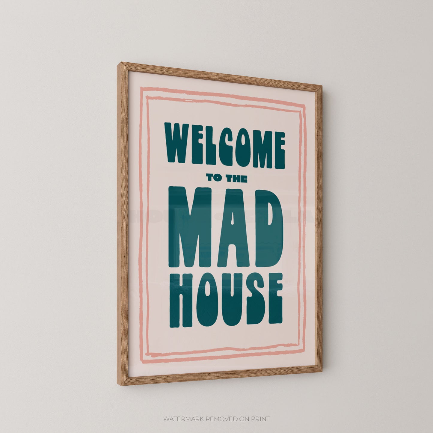 Framed wall art print with the text 'WELCOME TO THE MAD HOUSE' in teal lettering on a cream background, bordered by a thin pink line, in a light wood frame against a light gray wall.