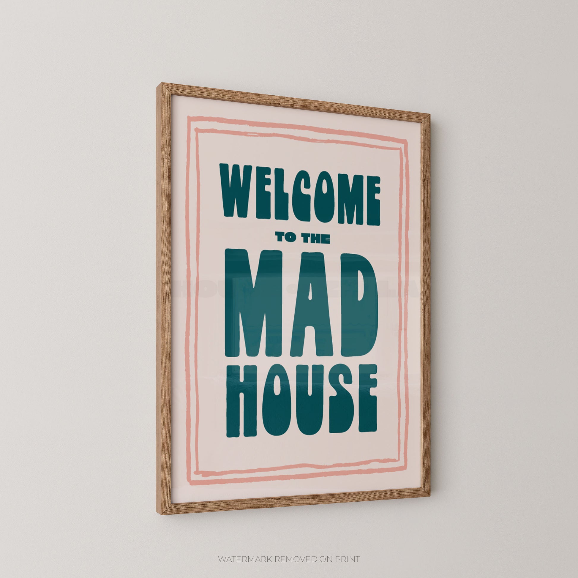 Framed wall art print with the text 'WELCOME TO THE MAD HOUSE' in teal lettering on a cream background, bordered by a thin pink line, in a light wood frame against a light gray wall.