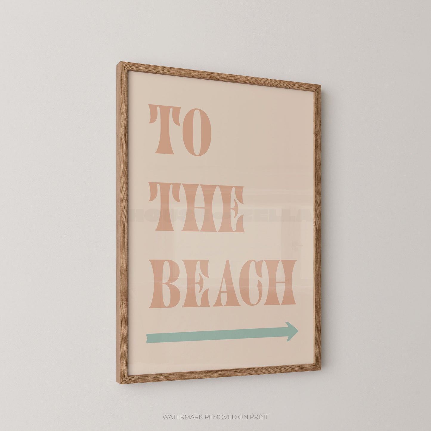Framed wall art print with the words “TO THE BEACH” in peach letters and a teal arrow below, shown hanging on a light gray wall.