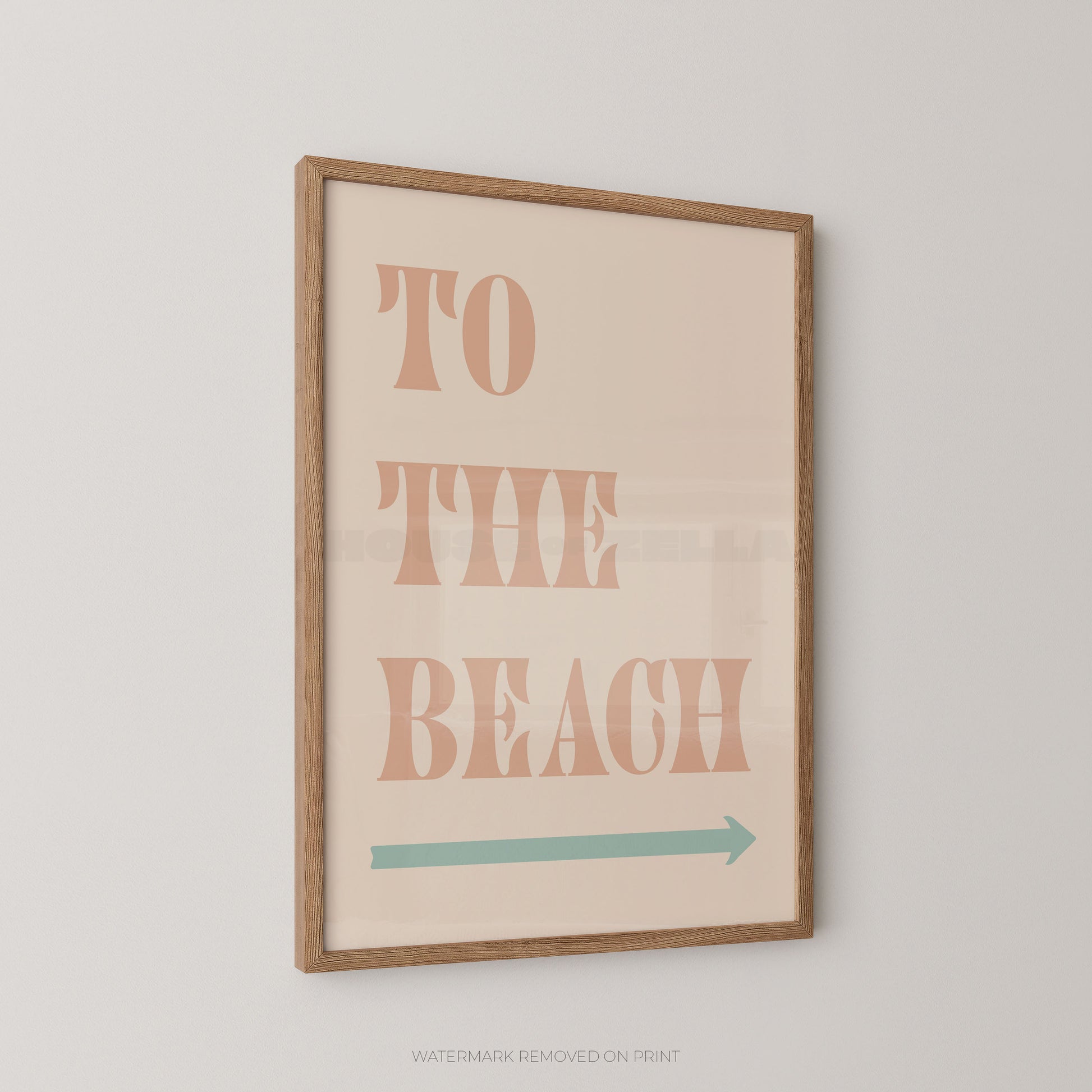Framed wall art print with the words “TO THE BEACH” in peach letters and a teal arrow below, shown hanging on a light gray wall.