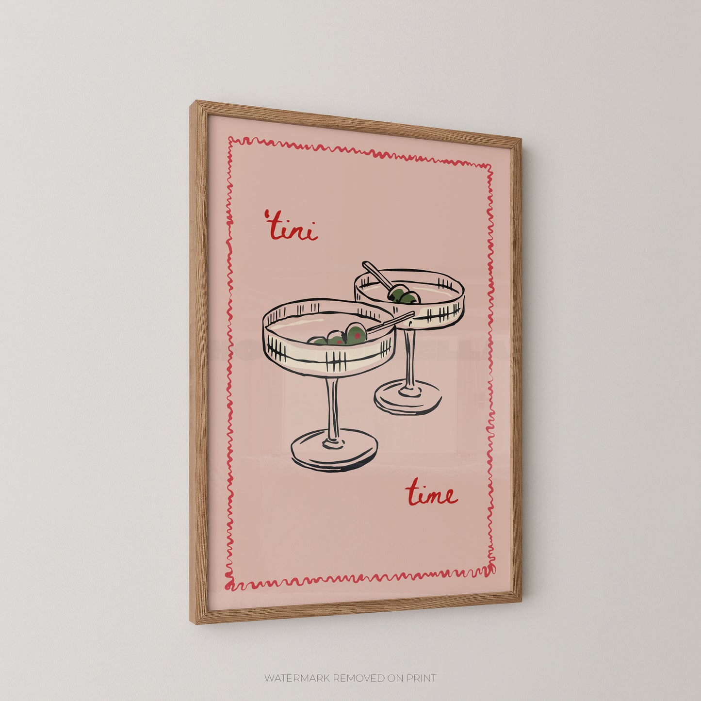 A framed art print featuring a retro illustration of two margarita glasses with olives, set against a dusty pink background with the words 'tini time' in red script. The print is housed in a light brown wooden frame and is displayed on a neutral light gray wall.