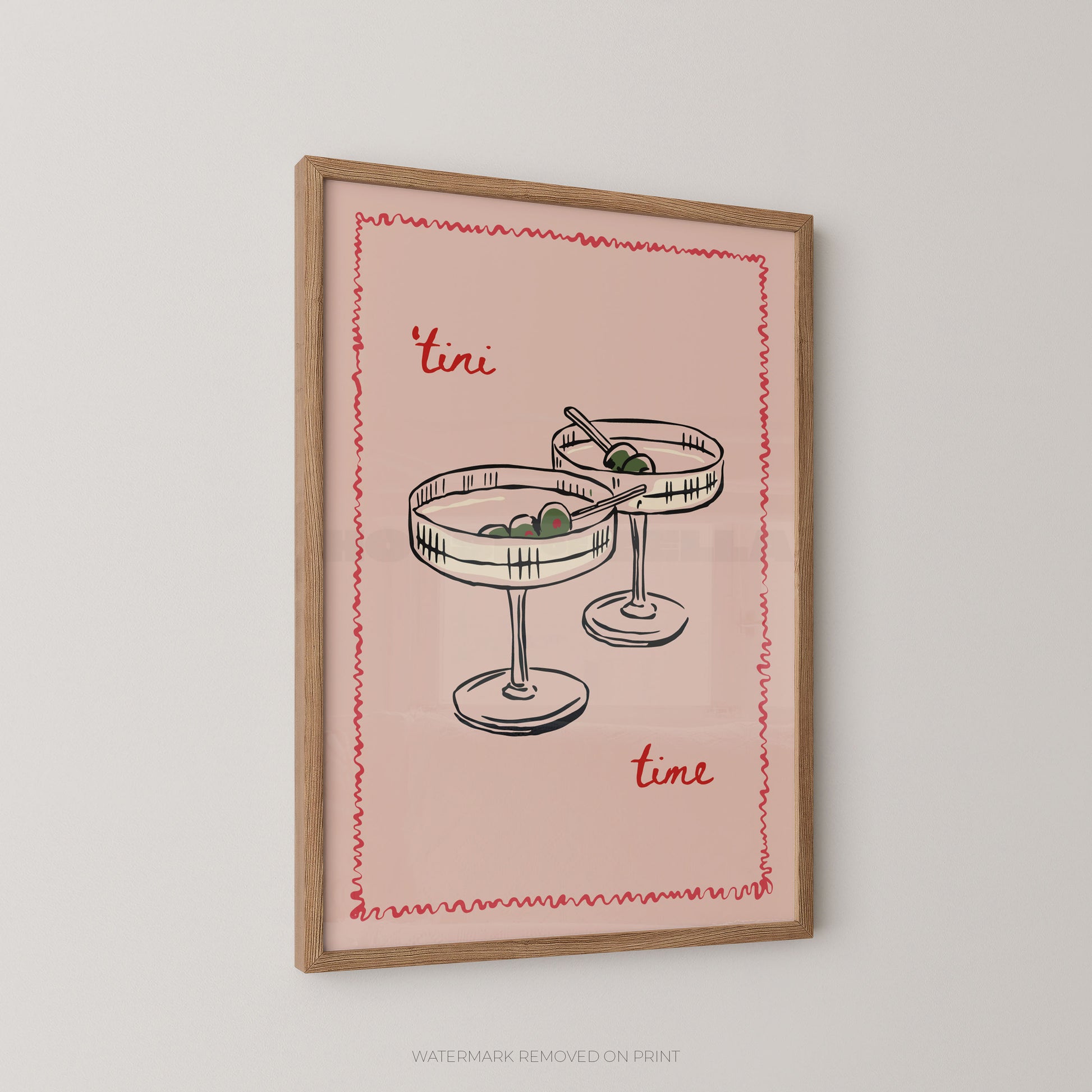 A framed art print featuring a retro illustration of two margarita glasses with olives, set against a dusty pink background with the words 'tini time' in red script. The print is housed in a light brown wooden frame and is displayed on a neutral light gray wall.