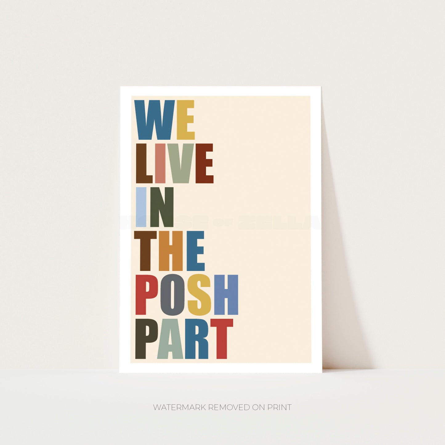 A vertical typographic art print with the phrase “WE LIVE IN THE POSH PART” in multicolored block letters on a warm cream background, shown leaning against a light gray wall.