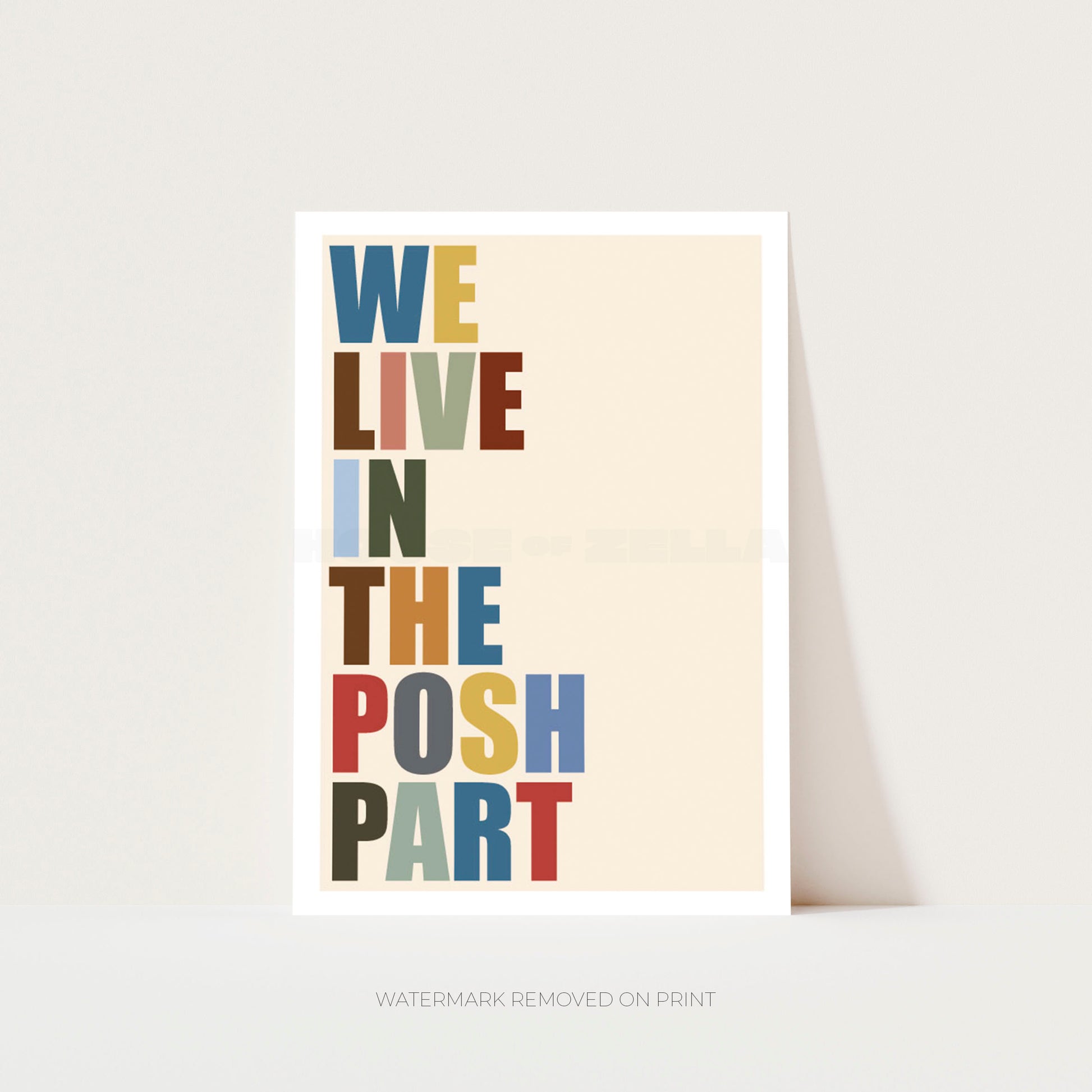 A vertical typographic art print with the phrase “WE LIVE IN THE POSH PART” in multicolored block letters on a warm cream background, shown leaning against a light gray wall.