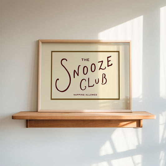 Framed artwork with 'The Snooze Club' text on a wooden shelf against a white wall.