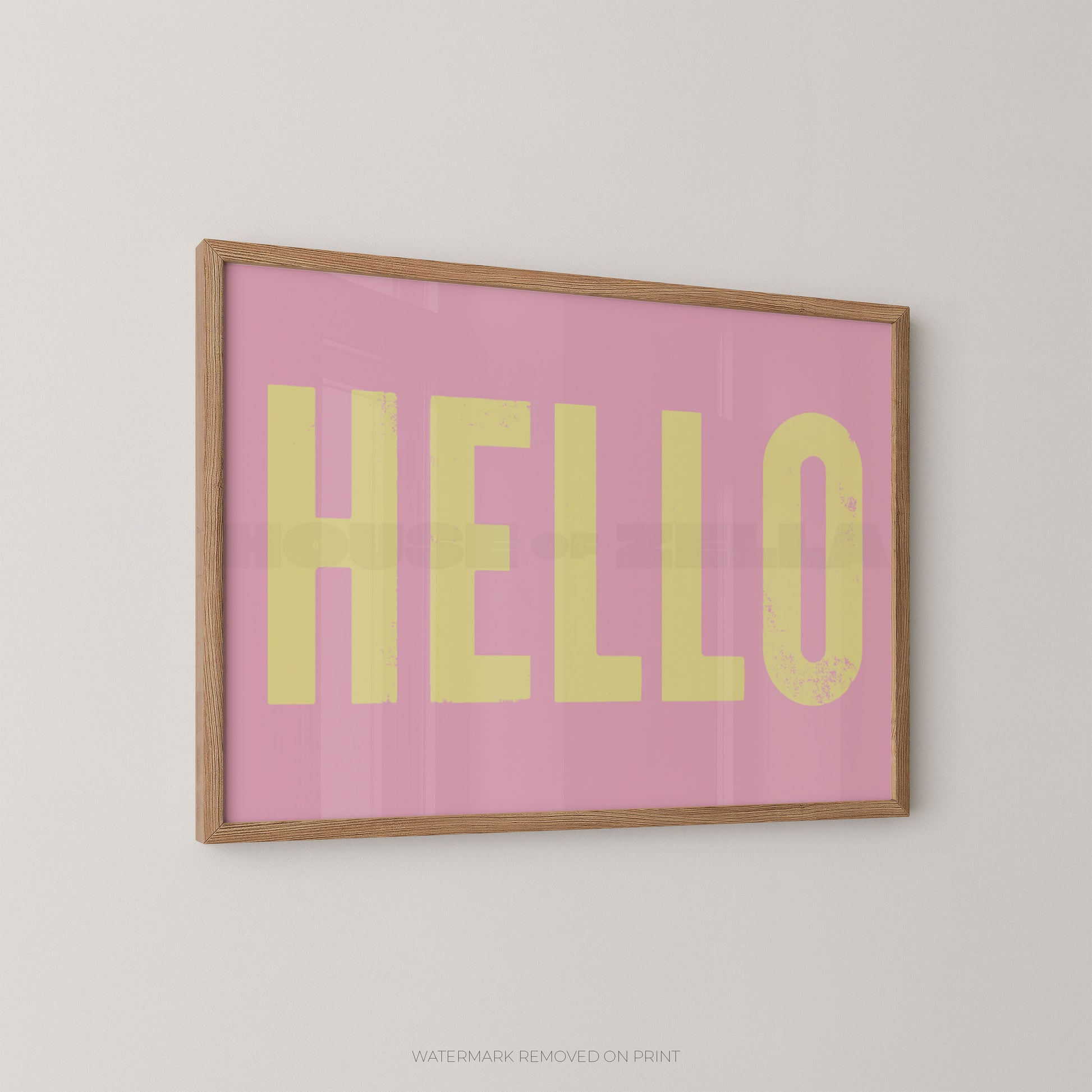 Framed wall art print with the word 'HELLO' in large yellow letters on a pink background, shown hanging on a light gray wall.
