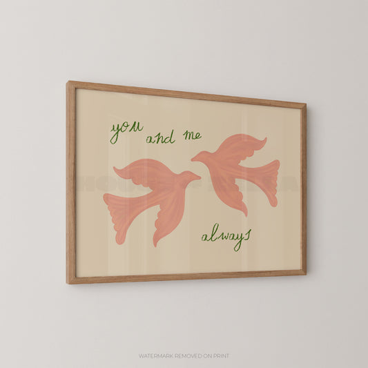 Framed wall art print with two pink flying birds and the words 'you and me always' in green script on a warm cream background, shown hanging on a light gray wall.