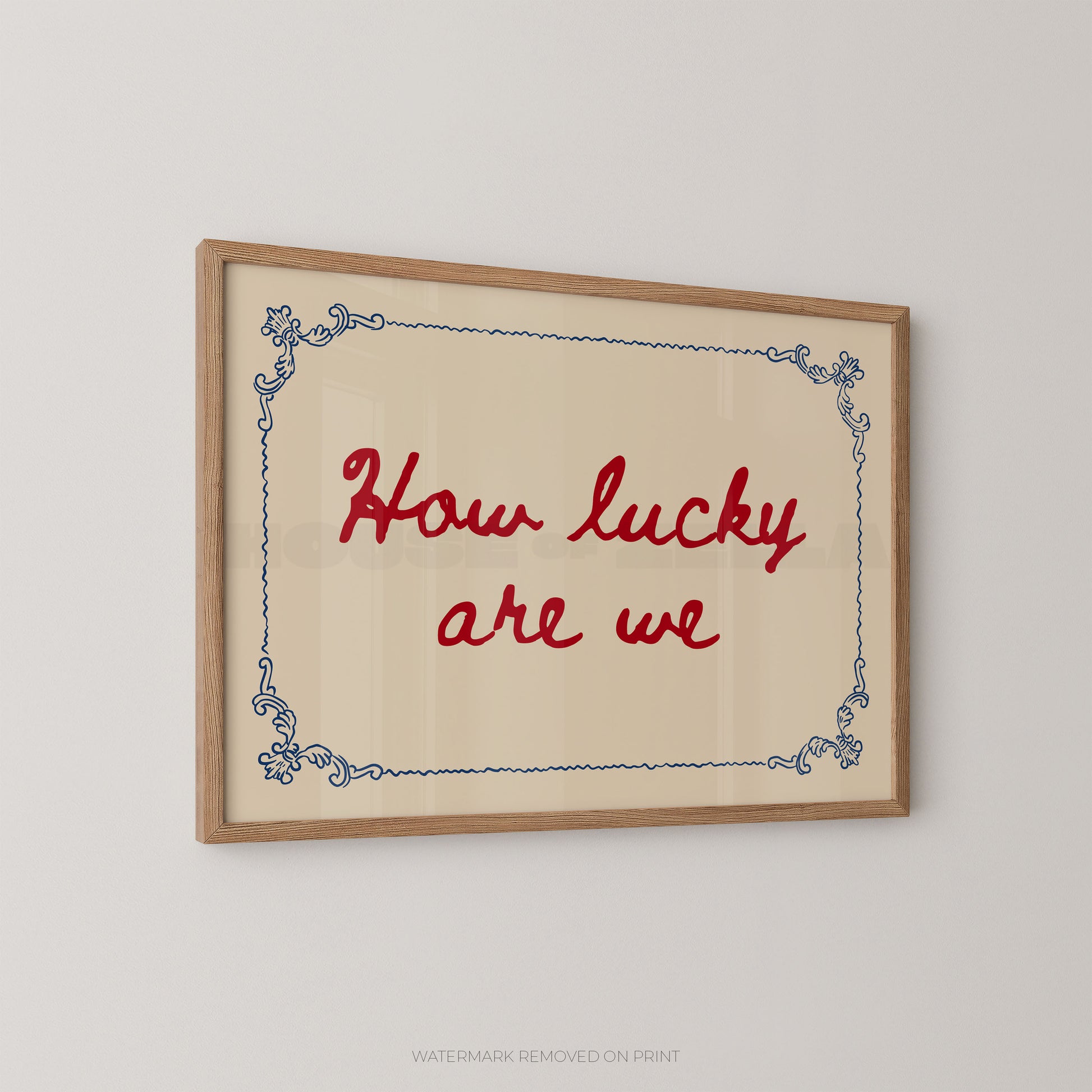 A framed typographic wall art print with the phrase “How lucky are we” in red script, set within a light wood frame and a cream background with a blue decorative border, shown hanging on a light gray wall.