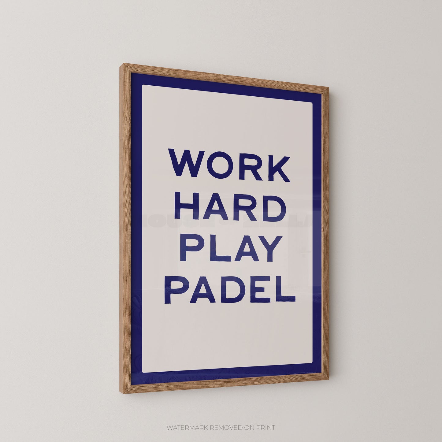 A framed typographic wall print with the text 'WORK HARD PLAY PADEL' in dark blue on a white background, bordered by a blue inner frame and a natural wood outer frame, shown hanging on a light gray wall.