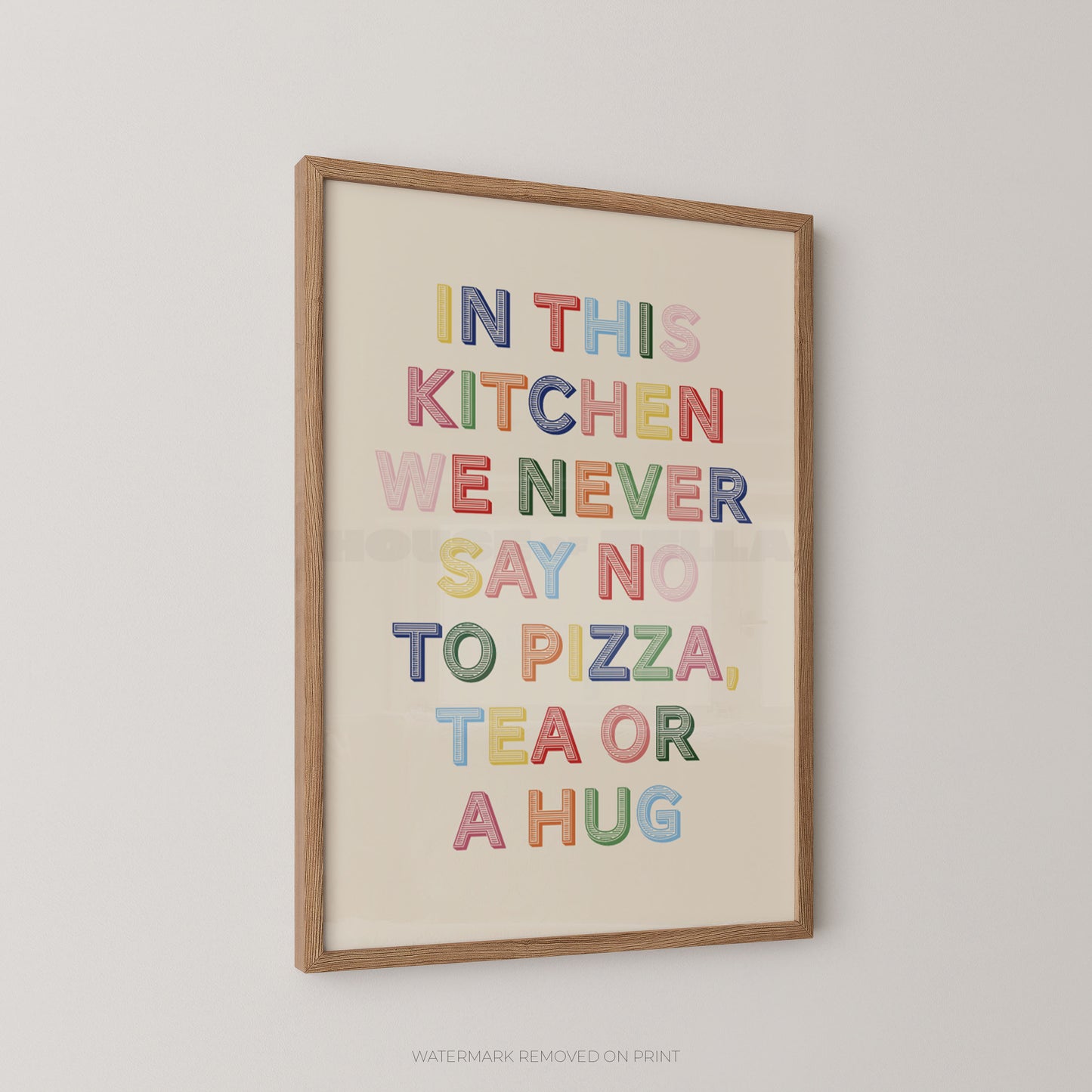 Framed wall art print with the phrase “IN THIS KITCHEN WE NEVER SAY NO TO PIZZA, TEA OR A HUG” in colorful raised letters on a cream background, shown hanging on a light gray wall.