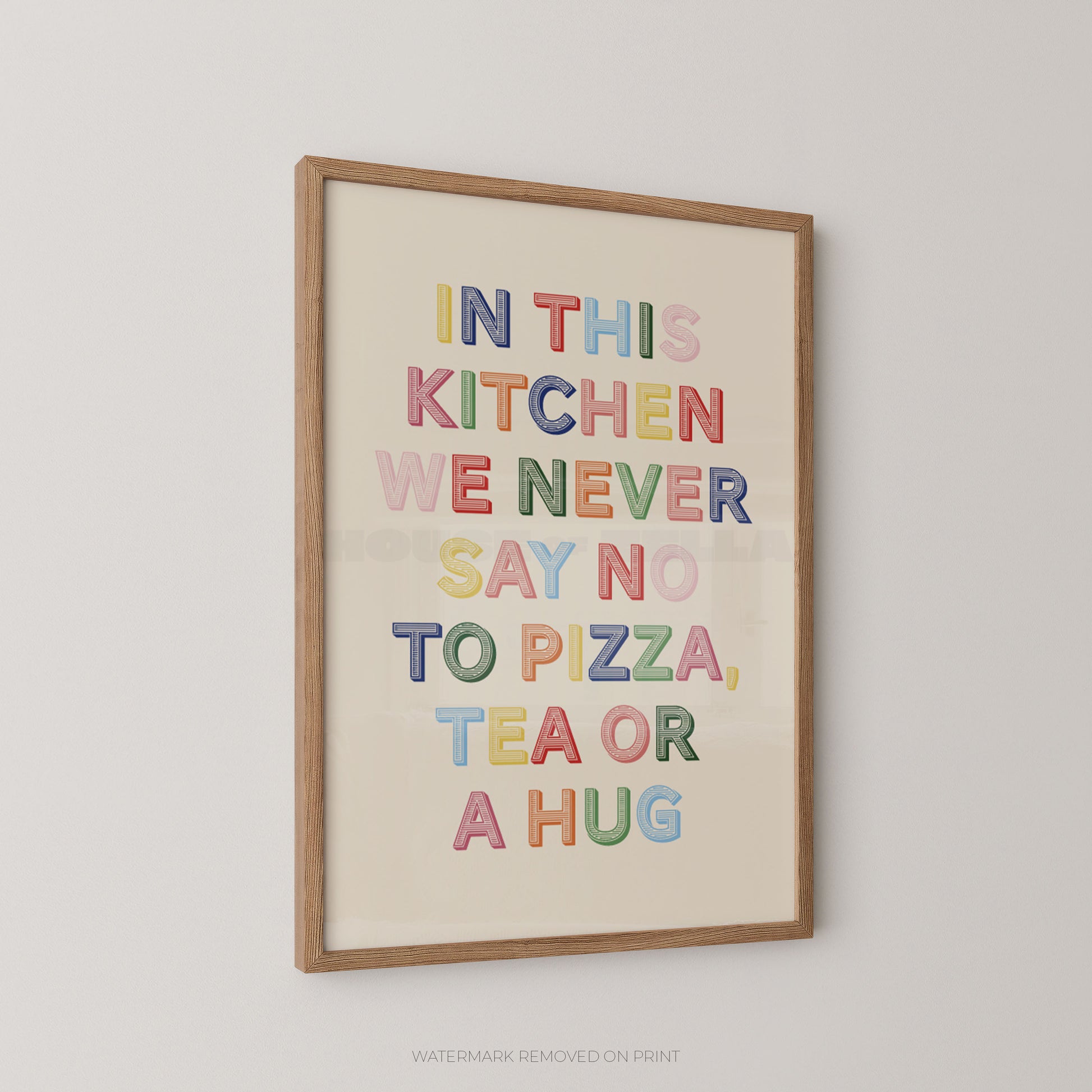 Framed wall art print with the phrase “IN THIS KITCHEN WE NEVER SAY NO TO PIZZA, TEA OR A HUG” in colorful raised letters on a cream background, shown hanging on a light gray wall.