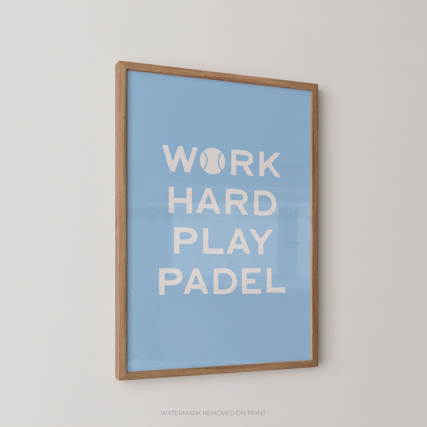 A framed wall art print with a light blue background and white text reading 'WORK HARD PLAY PADEL,' with a tennis ball icon replacing the letter O. The print is mounted in a natural wood frame and shown hanging on a light neutral wall.