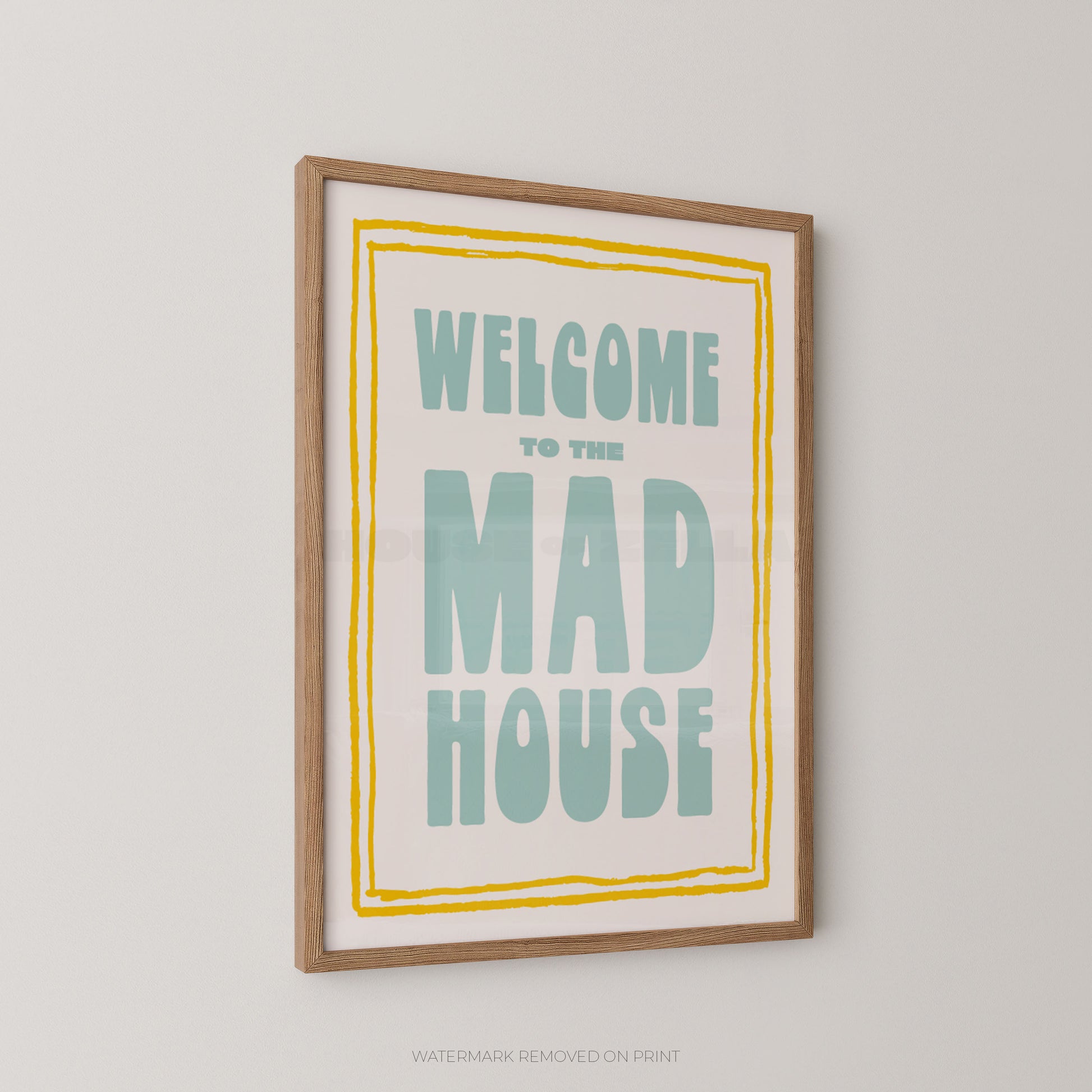Framed wall art print with the text 'WELCOME TO THE MAD HOUSE' in bold pale blue lettering on a cream background, with a yellow border enclosed in a light wood frame and shown hanging on a light gray wall.