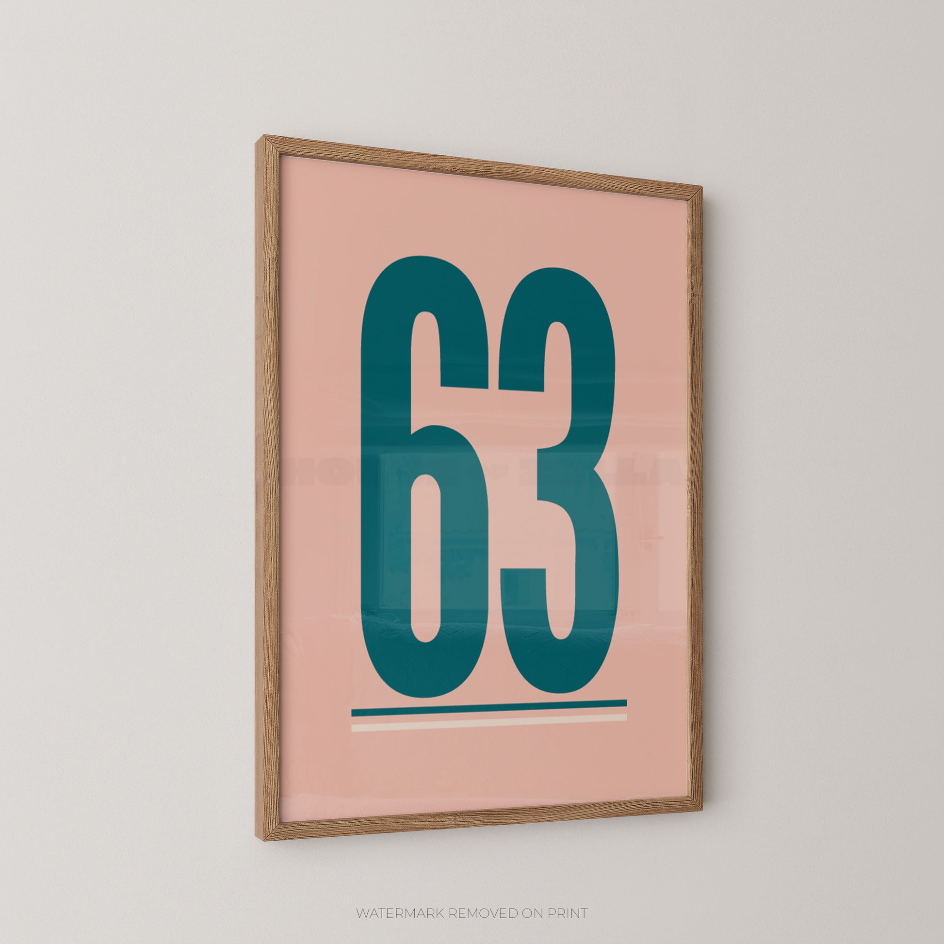 Framed minimalist wall art featuring the number 63 in teal on a blush pink background, hung on a light gray wall.
