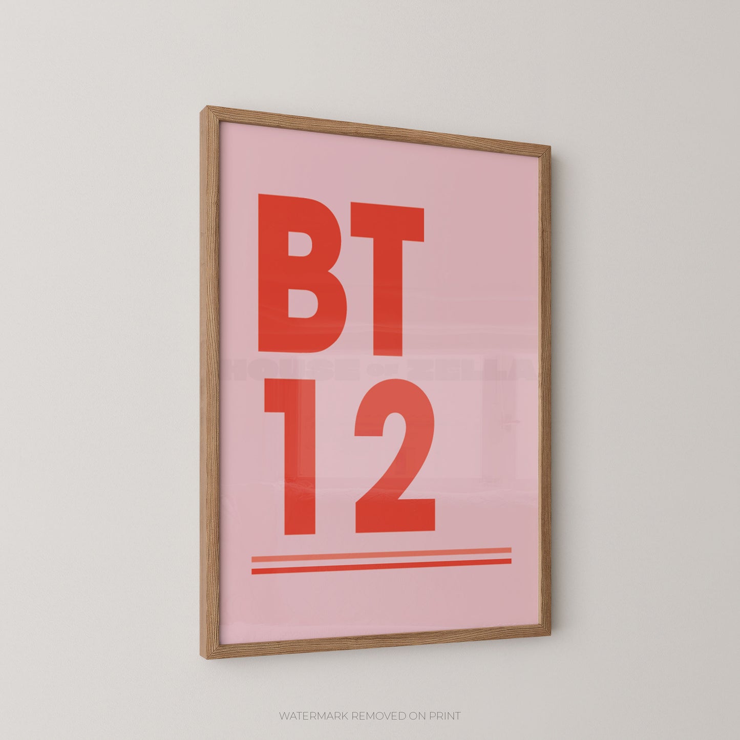 A framed typographic wall art print with a light pink background, bold red text reading 'BT 12', and two thin red horizontal lines, shown hanging on a light gray wall.