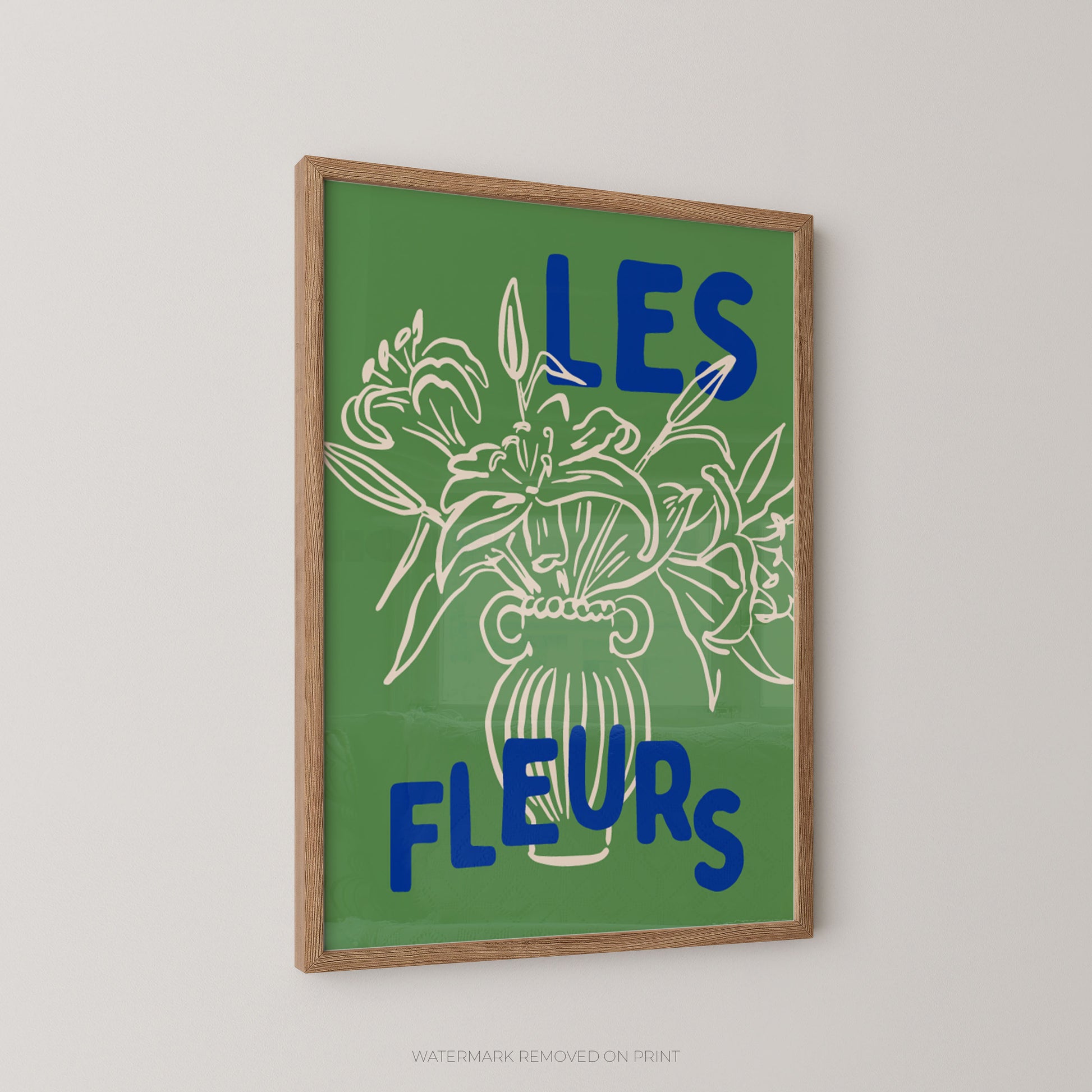A framed art print featuring a green background with a white line drawing of flowers in a vase and the blue French text 'LES FLEURS' on a light gray wall.