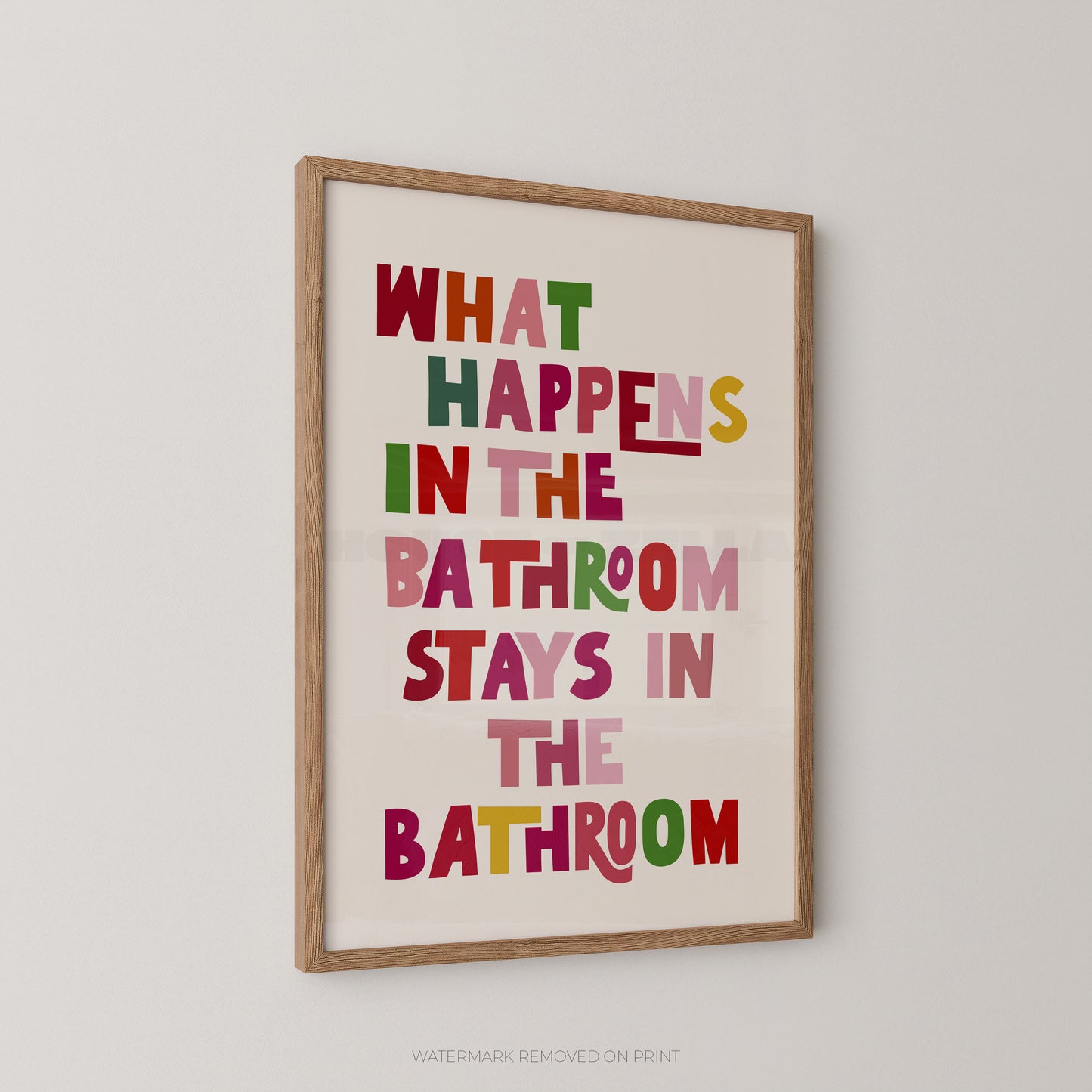 Funny bathroom wall art print in a wood frame – playful multicoloured chunky typography on a cream background reading "What Happens In The Bathroom Stays In The Bathroom"