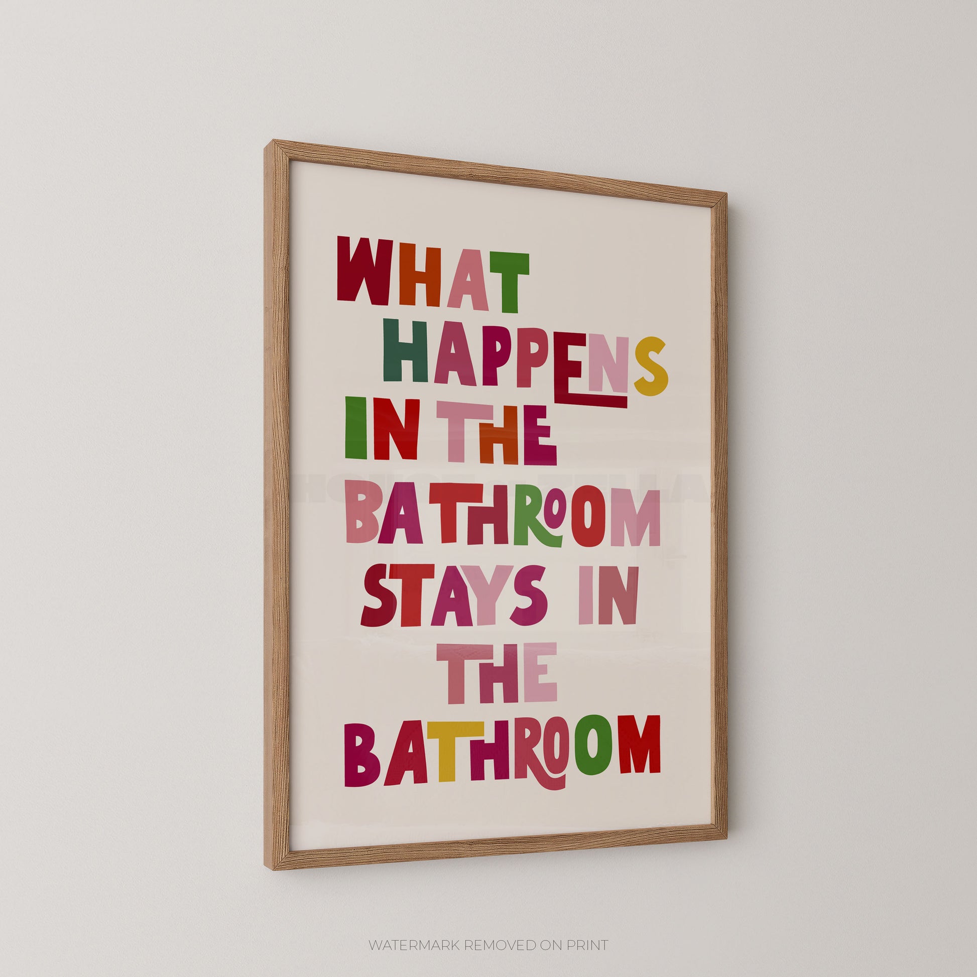 Funny bathroom wall art print in a wood frame – playful multicoloured chunky typography on a cream background reading "What Happens In The Bathroom Stays In The Bathroom"