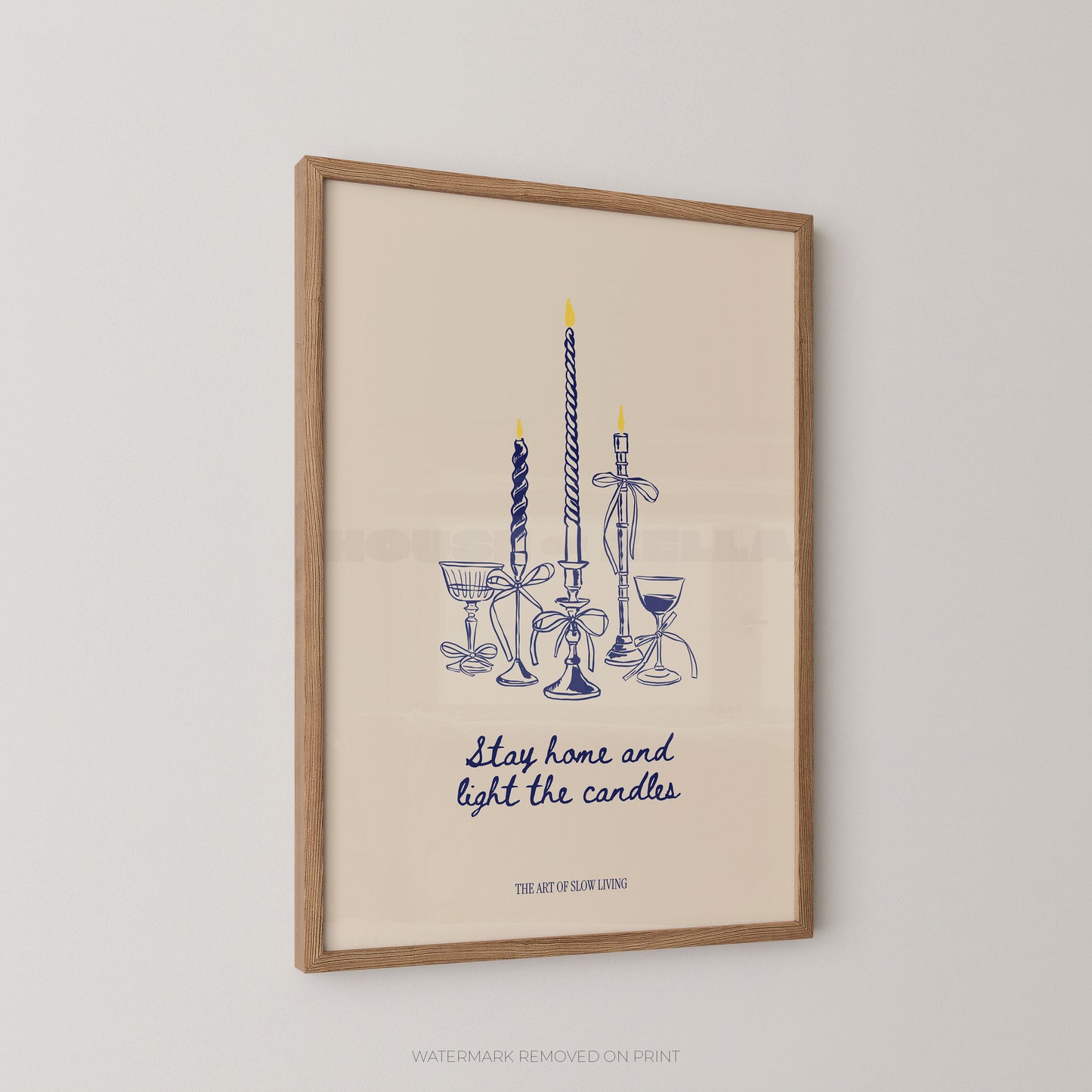 Framed wall art print featuring a blue line drawing of candles and goblets with the text 'Stay home and light the candles' and 'THE ART OF SLOW LIVING on a warm cream background, shown hanging on a light wall.
