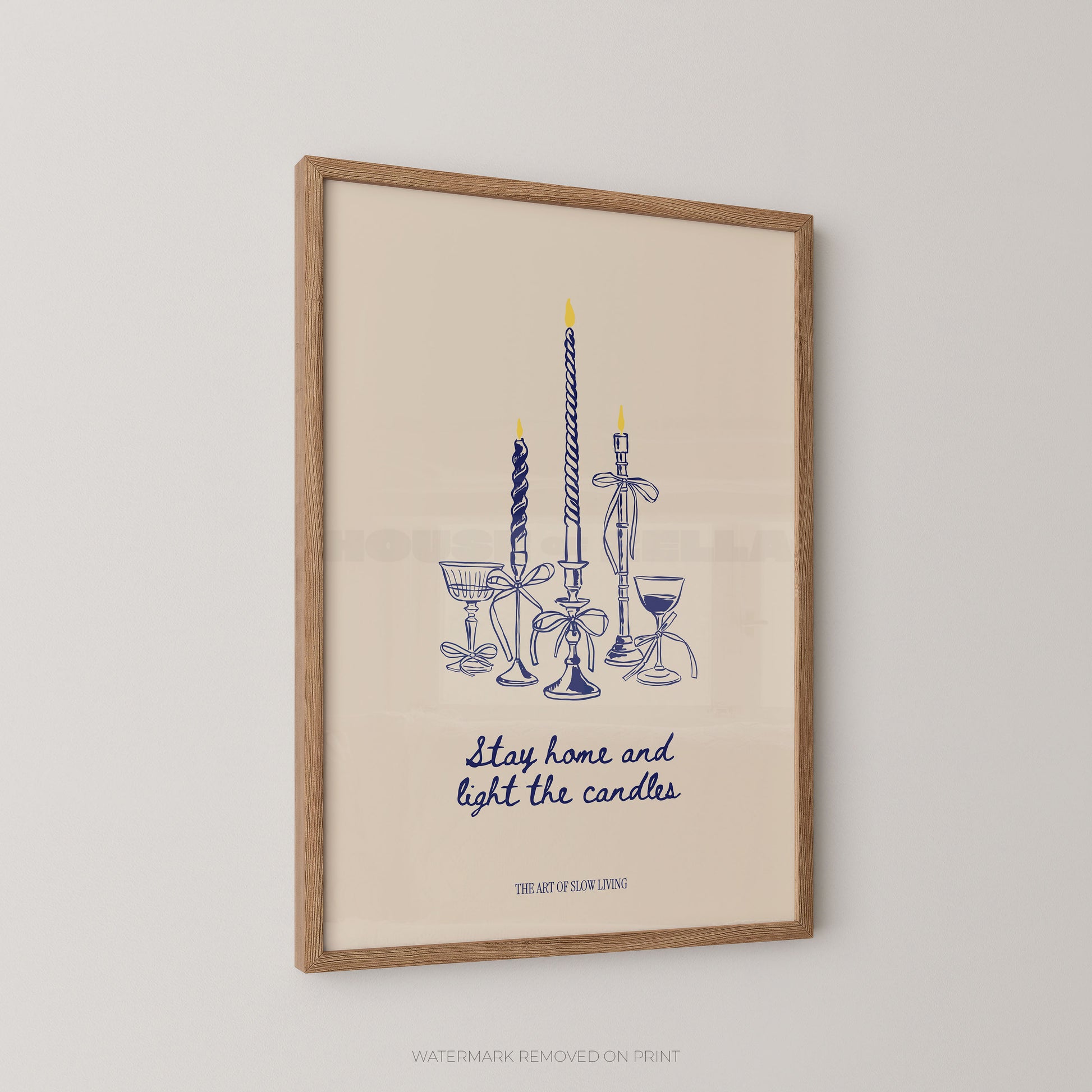 Framed wall art print featuring a blue line drawing of candles and goblets with the text 'Stay home and light the candles' and 'THE ART OF SLOW LIVING on a warm cream background, shown hanging on a light wall.