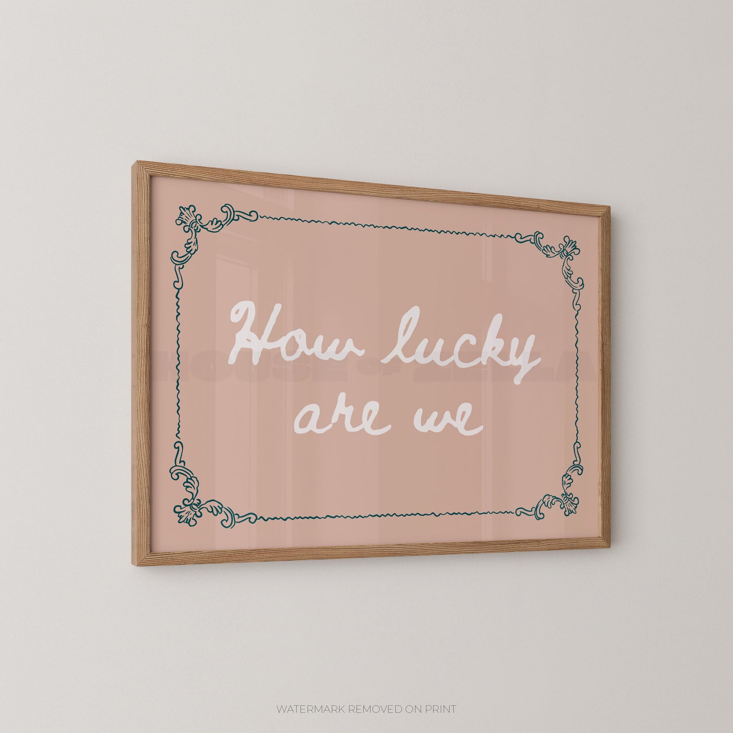 A framed wall art print with the phrase “How lucky are we” in white script on a soft blush pink background, bordered by a light wood frame and a teal decorative corner pattern, shown hanging on a light gray wall.