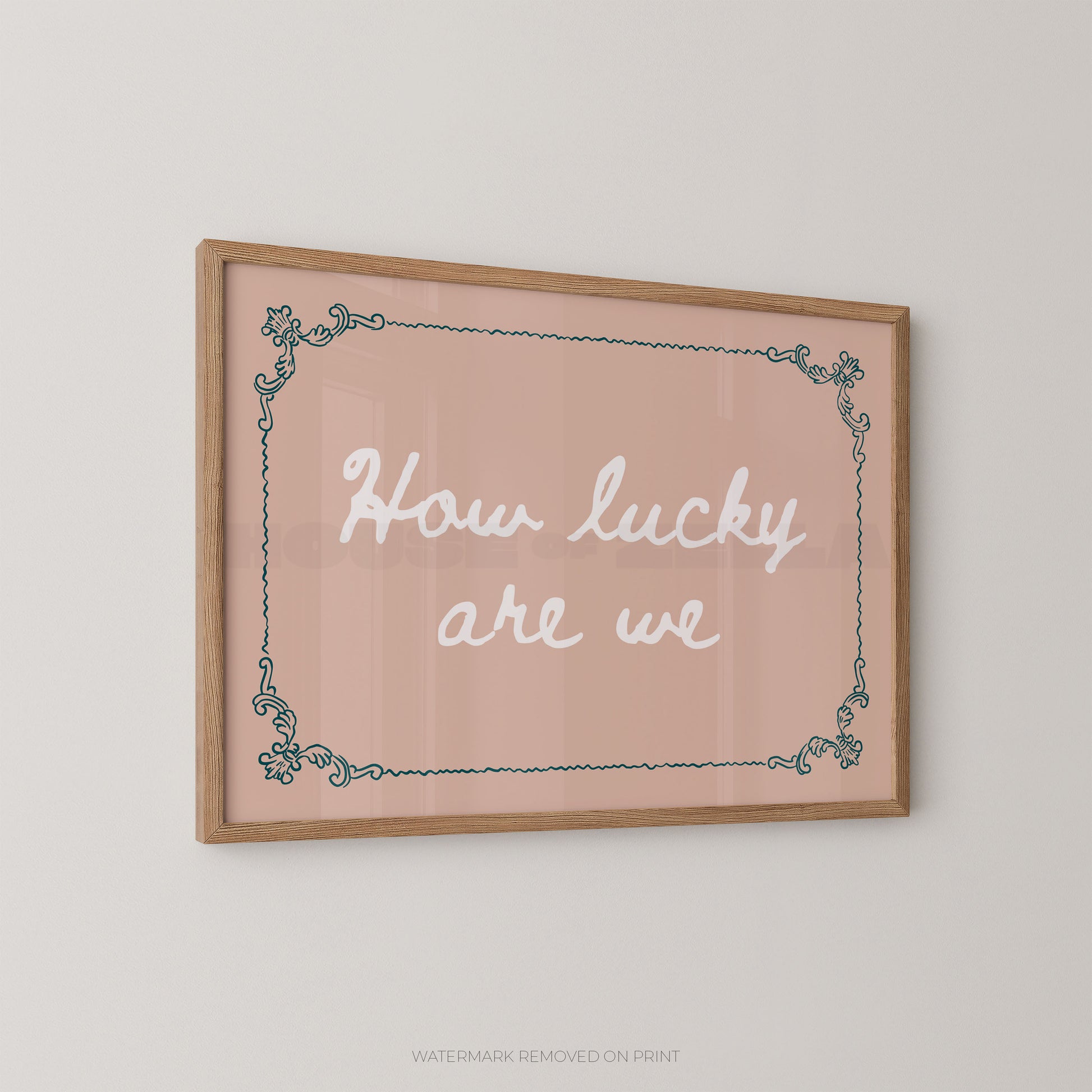 A framed wall art print with the phrase “How lucky are we” in white script on a soft blush pink background, bordered by a light wood frame and a teal decorative corner pattern, shown hanging on a light gray wall.