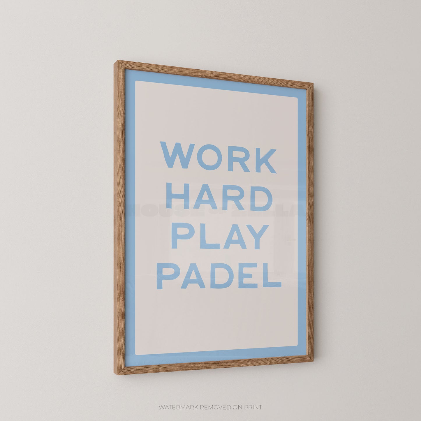 A framed typographic wall print with the words “WORK HARD PLAY PADEL” in light blue on a white background, bordered by a light blue inner border and a natural wood frame, shown hanging on a light gray wall.