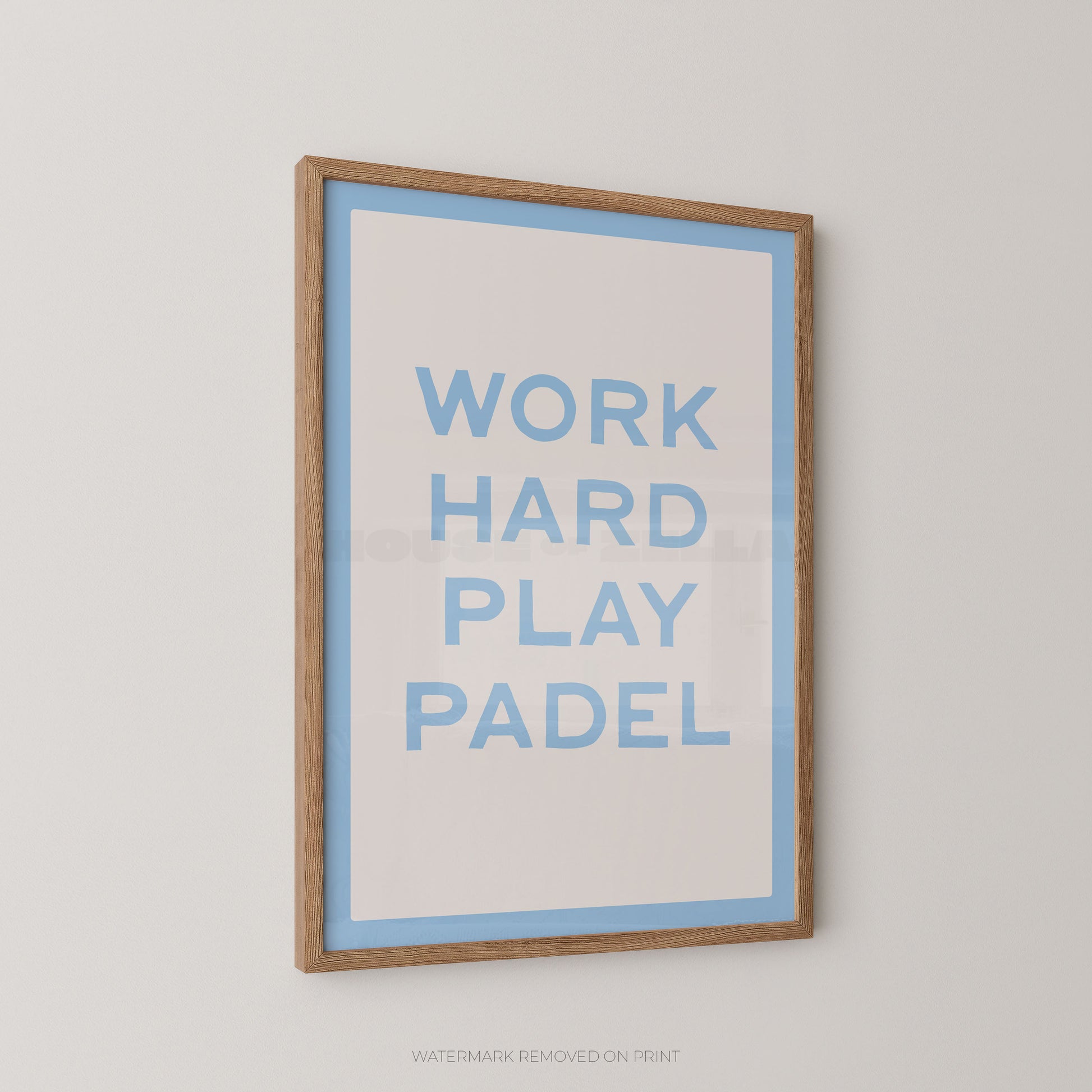 A framed typographic wall print with the words “WORK HARD PLAY PADEL” in light blue on a white background, bordered by a light blue inner border and a natural wood frame, shown hanging on a light gray wall.
