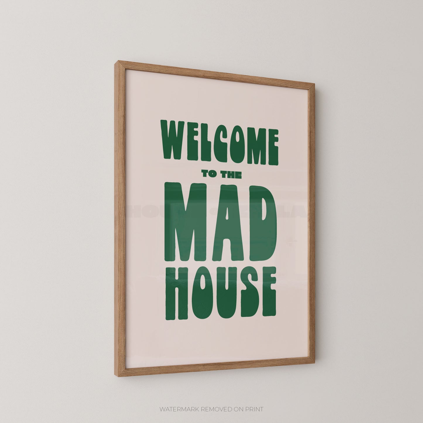 Framed wall art print with the phrase “WELCOME TO THE MAD HOUSE” in large green letters on a pale pink background, shown hanging on a light gray wall.