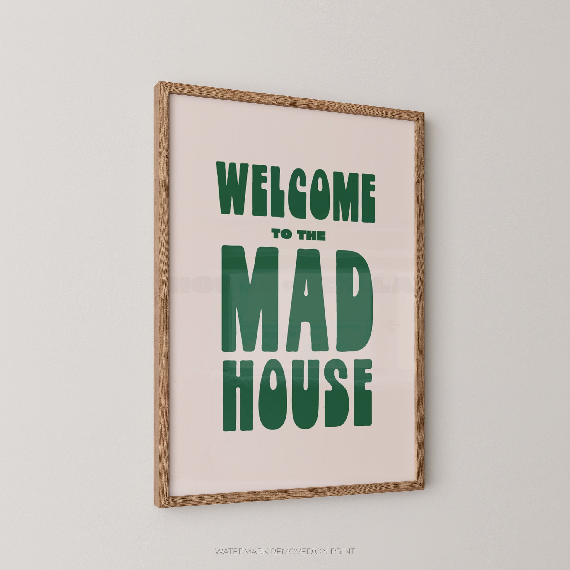 Framed wall art print with the phrase “WELCOME TO THE MAD HOUSE” in large green letters on a pale pink background, shown hanging on a light gray wall.
