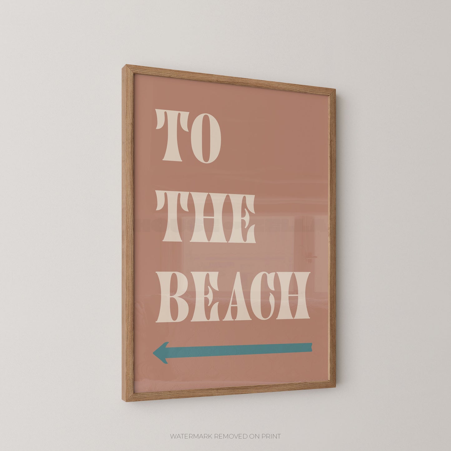 Framed wall art print with the words “TO THE BEACH” in large cream letters on a dusty pink background, plus a teal left-pointing arrow, shown hanging on a light neutral wall.